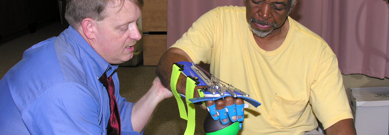 Dr. Jeffrey Kinsella-Shaw holds the arm of a patient who is wearing an assistive device on his arm and hand while lifting a ball.
